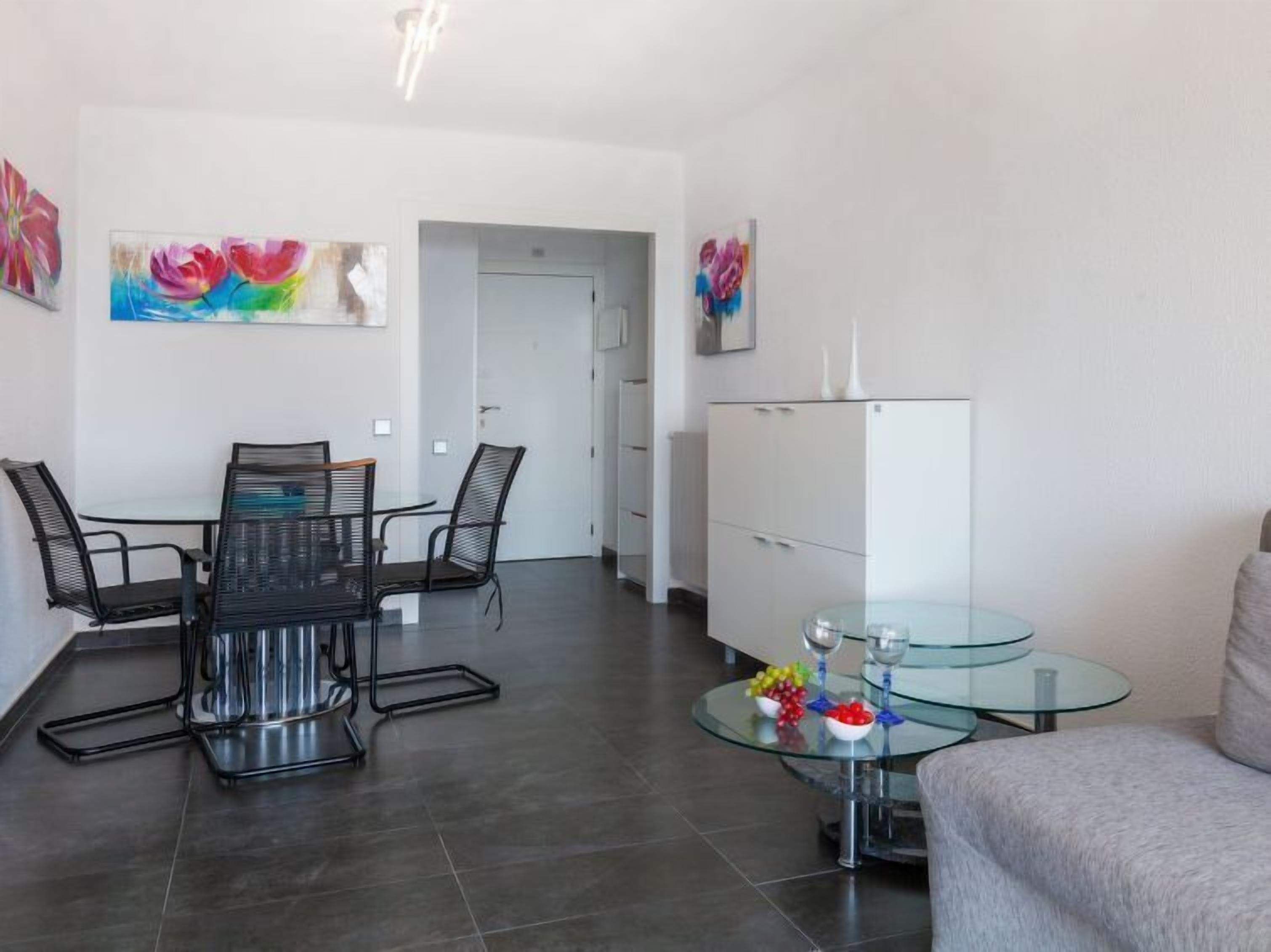 Una sala moderna con una mesa de comedor de vidrio, sillas de metal negro, una mesa de centro de vidrio con objetos decorativos, un aparador blanco y pinturas coloridas en las paredes.