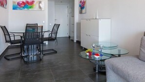 Una sala moderna con una mesa de comedor de vidrio, sillas de metal negro, una mesa de centro de vidrio con objetos decorativos, un aparador blanco y pinturas coloridas en las paredes.