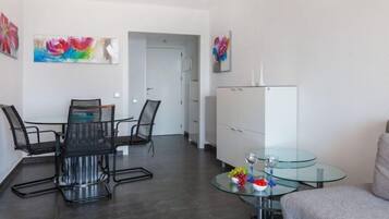 Una sala moderna con una mesa de comedor de vidrio, sillas de metal negro, una mesa de centro de vidrio con objetos decorativos, un aparador blanco y pinturas coloridas en las paredes.