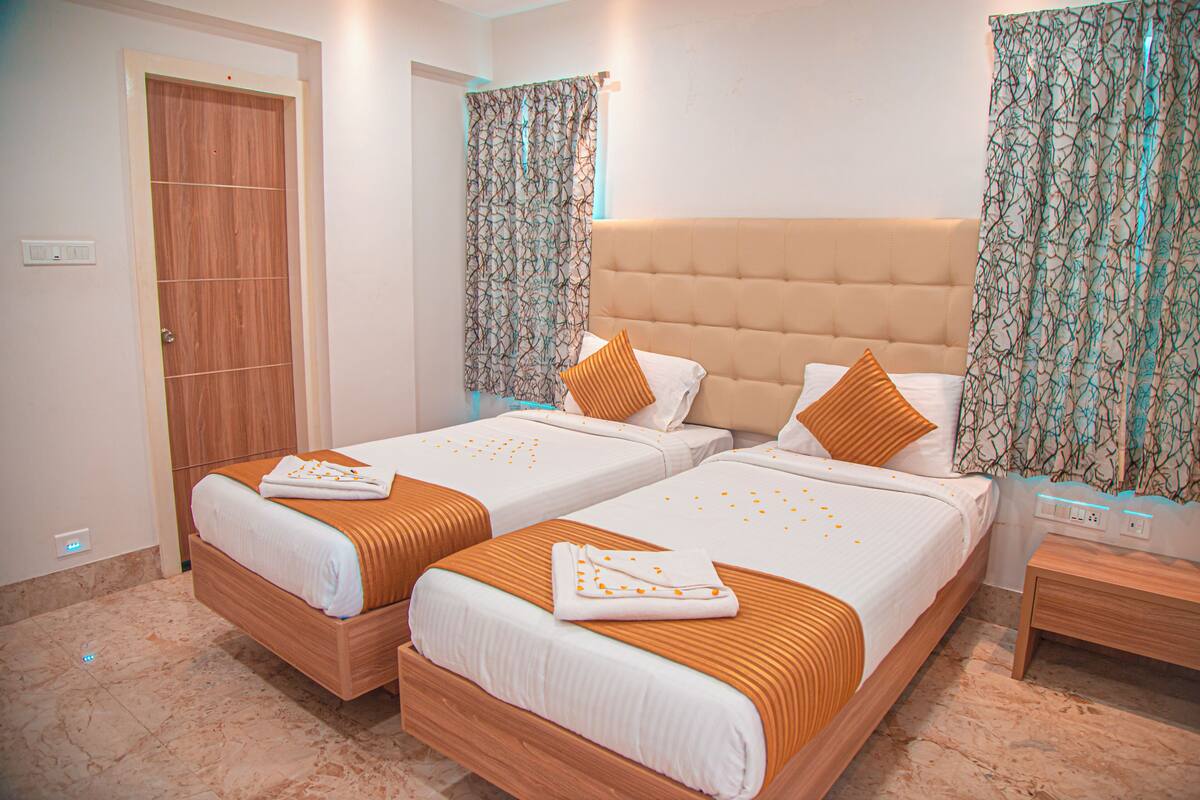 A hotel room with two beds, a wooden door, a bathroom with a shower curtain, and a nightstand with a lamp.