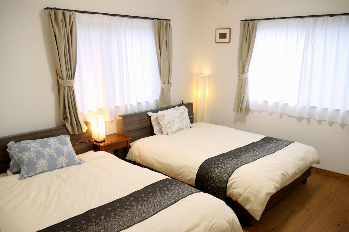 A hotel room with two single beds, wooden headboards, a nightstand with a lamp, and curtains on the windows.