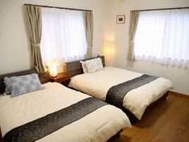 Habitación de hotel con dos camas individuales, cabeceros de madera, una mesita de noche con lámpara y cortinas en las ventanas.