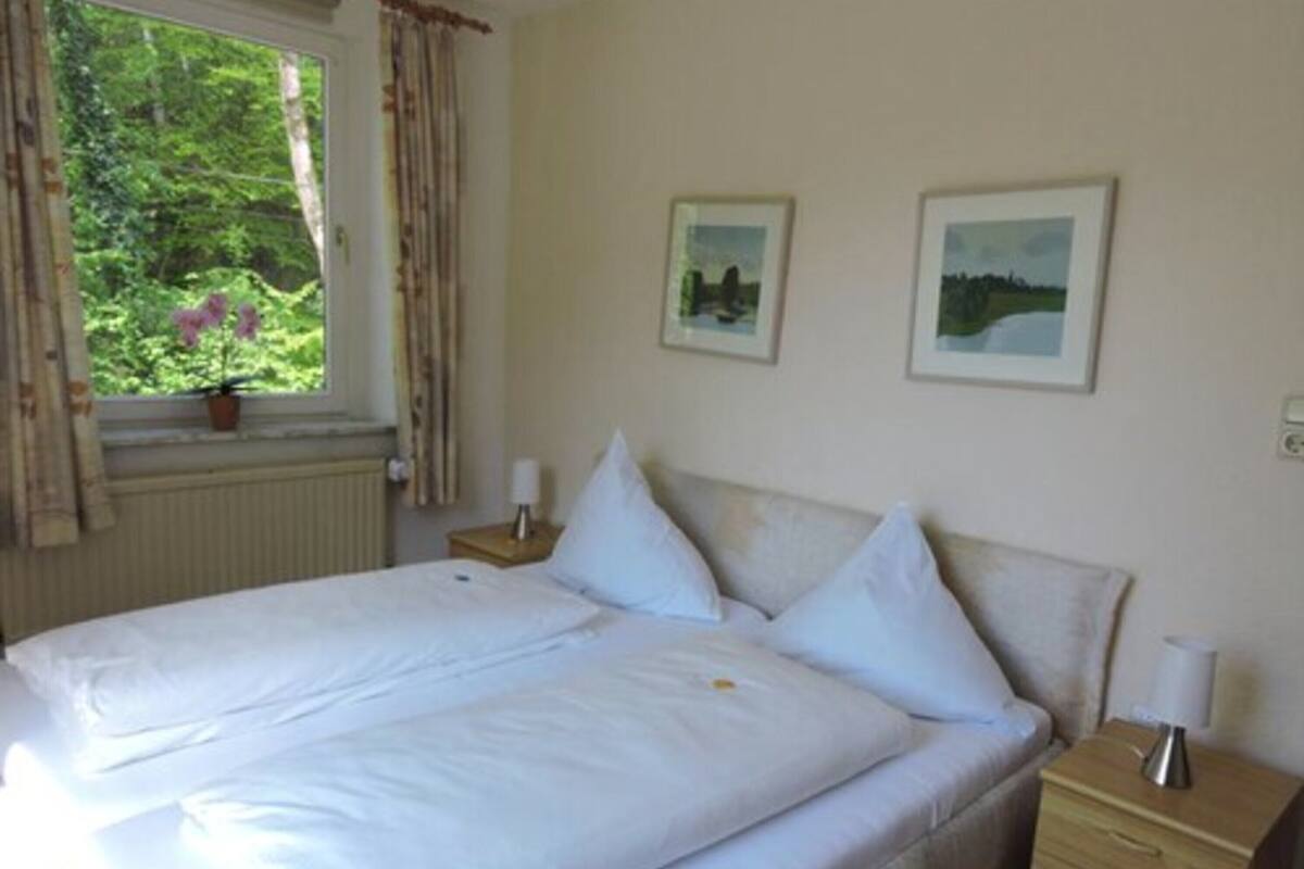 A double bed with white linens, a bedside table with a lamp, and two framed pictures on the wall.