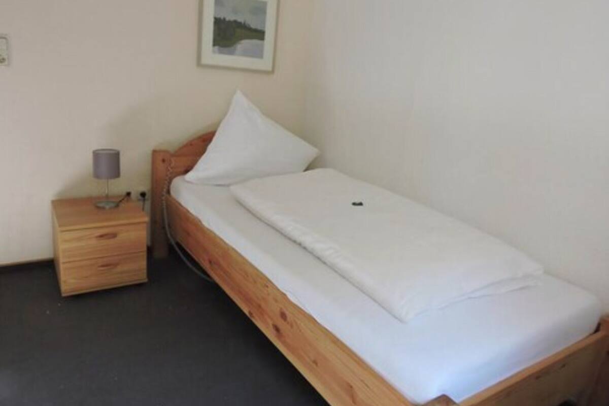 A small, single-bed room with a wooden bed frame, a bedside table with a lamp, and a framed picture on the wall.