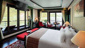 A hotel room with a large bed, a desk with a chair, a TV, and a view of a lake. - Orchid Premium Cruises (Hai Phong)