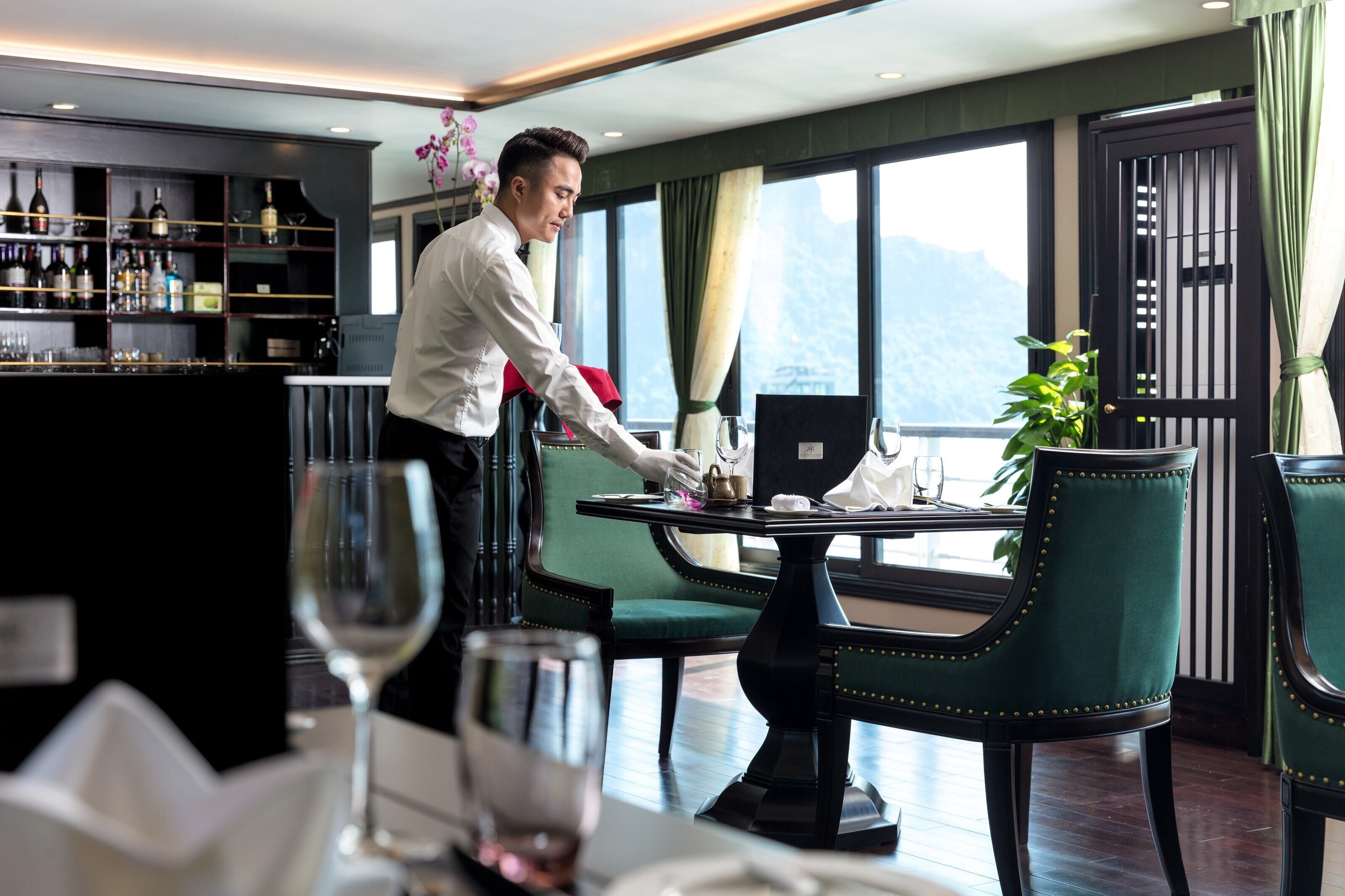 A restaurant with a bar area, green upholstered chairs, and a view of mountains through large windows.