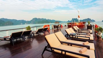 A deck with lounge chairs and tables, a flag, and a view of the water and mountains.