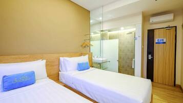 Habitación de hotel con dos camas, cabecero de madera, mampara de ducha de vidrio, puerta de madera y unidad de aire acondicionado.