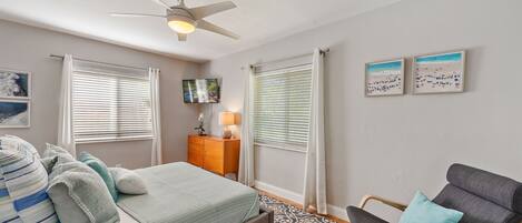 Une chambre à coucher comprenant un lit, une chaise, un ventilateur de plafond et deux fenêtres équipées de stores.
