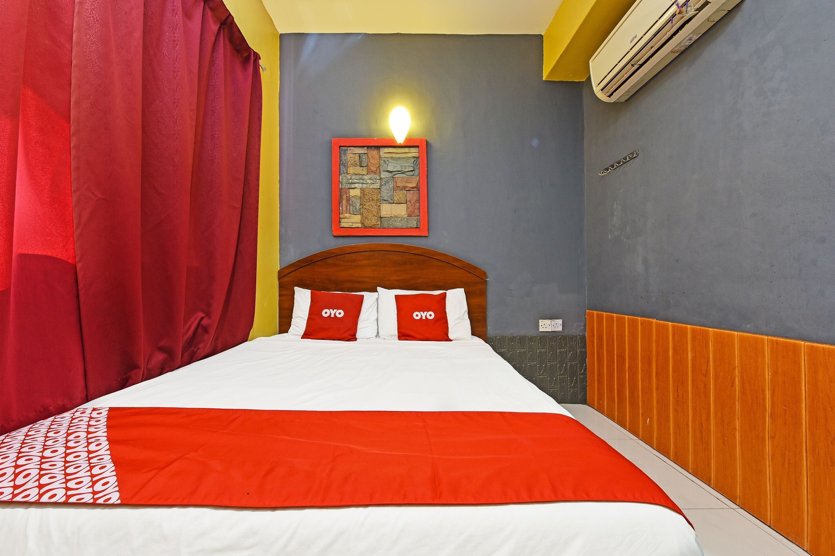 A hotel room with a bed, red curtains, a wall-mounted artwork, and an air conditioning unit.