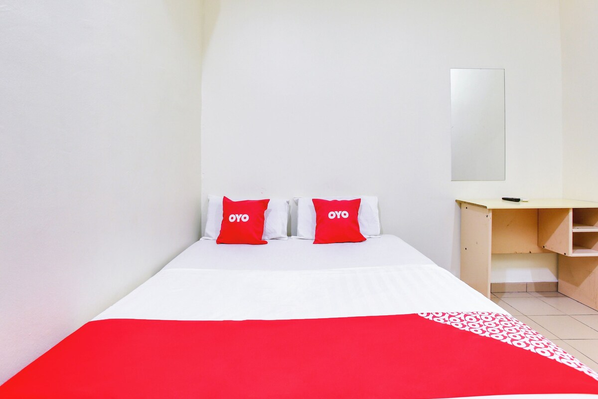 Una habitación de hotel con una cama, dos almohadas rojas con la marca 'OYO', un escritorio de madera y un espejo en la pared.