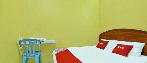 A room with a bed, a wooden headboard, a blue plastic chair, and a small desk with a telephone.