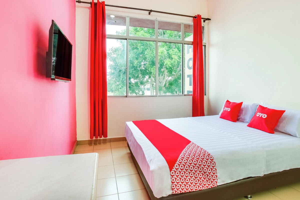 Habitación de hotel con cama, televisor montado en la pared, ventana con cortinas rojas y vista a zonas verdes.