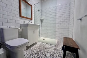 Une salle de bain avec des murs recouverts de carreaux blancs, des toilettes blanches, un lavabo et un espace douche.