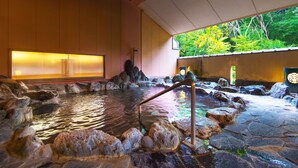 Un onsen japonais traditionnel, avec un bain en pierre entouré de rochers, un banc en bois et une vue sur une végétation luxuriante.