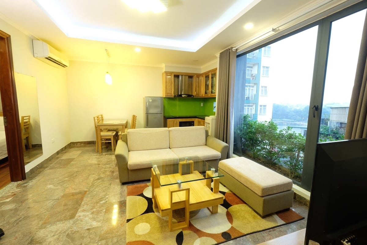 A modern living room with a sofa, a glass coffee table, and a TV. There's a kitchen area with wooden cabinets and a green backsplash.