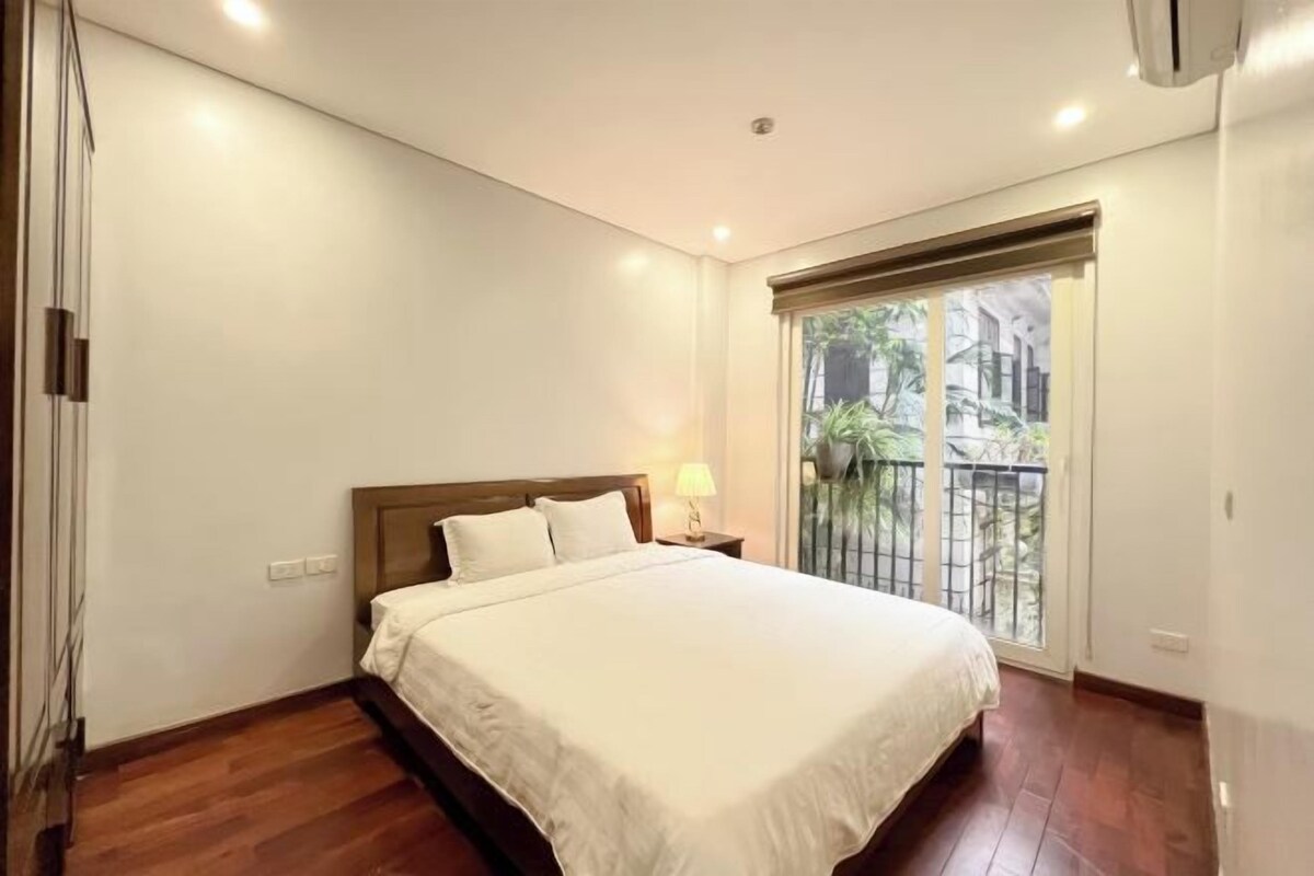 A bedroom with a bed, a nightstand, a window with a view, and a door.