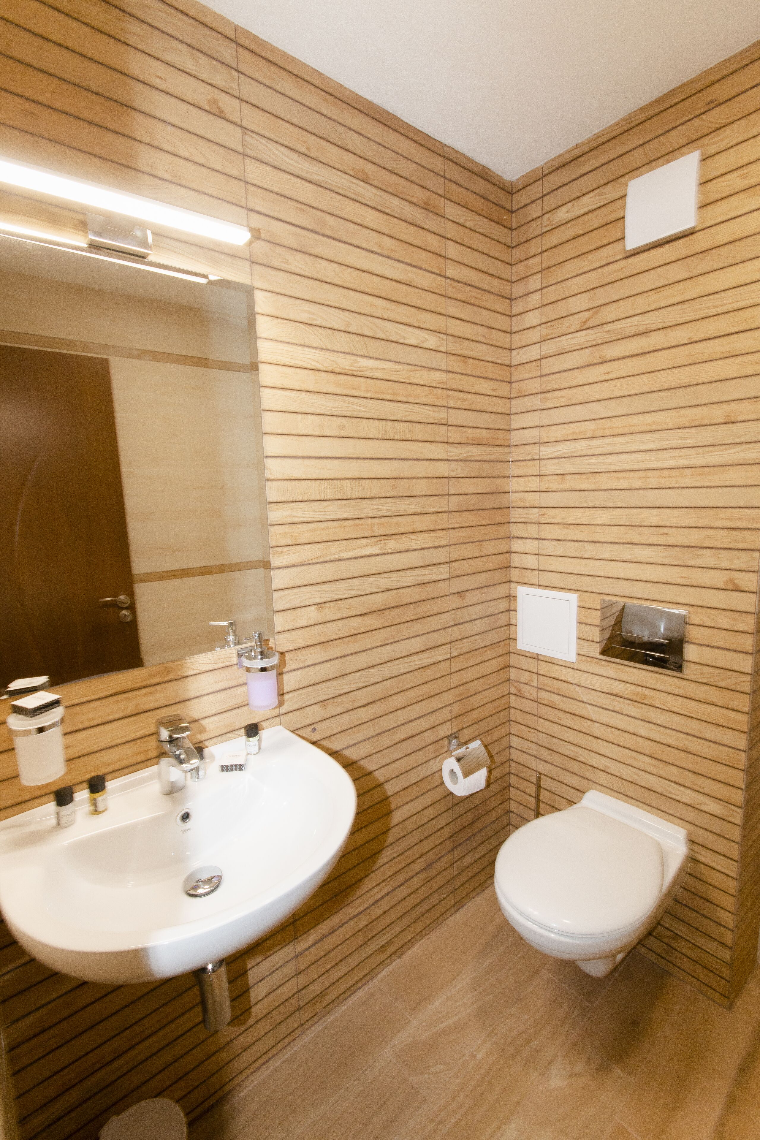 Une salle de bain avec des murs recouverts de panneaux en bois, un lavabo blanc, des toilettes et un miroir.