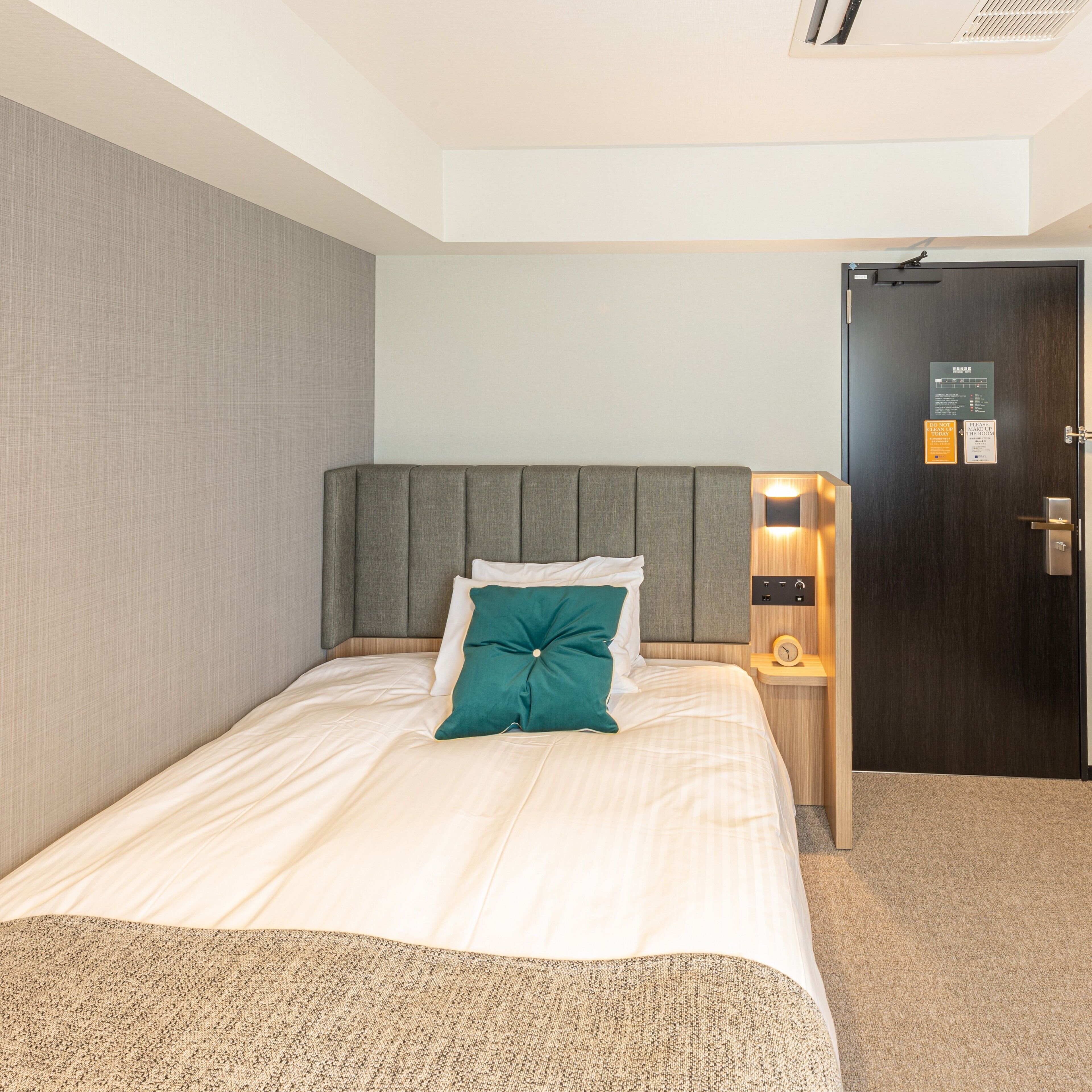 A hotel room with a bed, a nightstand, a wall-mounted light, and a door with a sign.