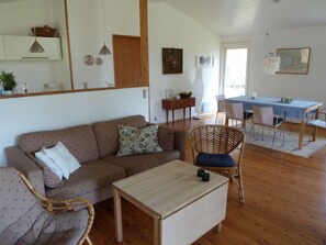 A living room with a sofa, a coffee table, a wicker chair, a dining table with chairs, and a kitchen area in the background. - Lovely cottage in Oddsherred, close to child-friendly beach (Højby)