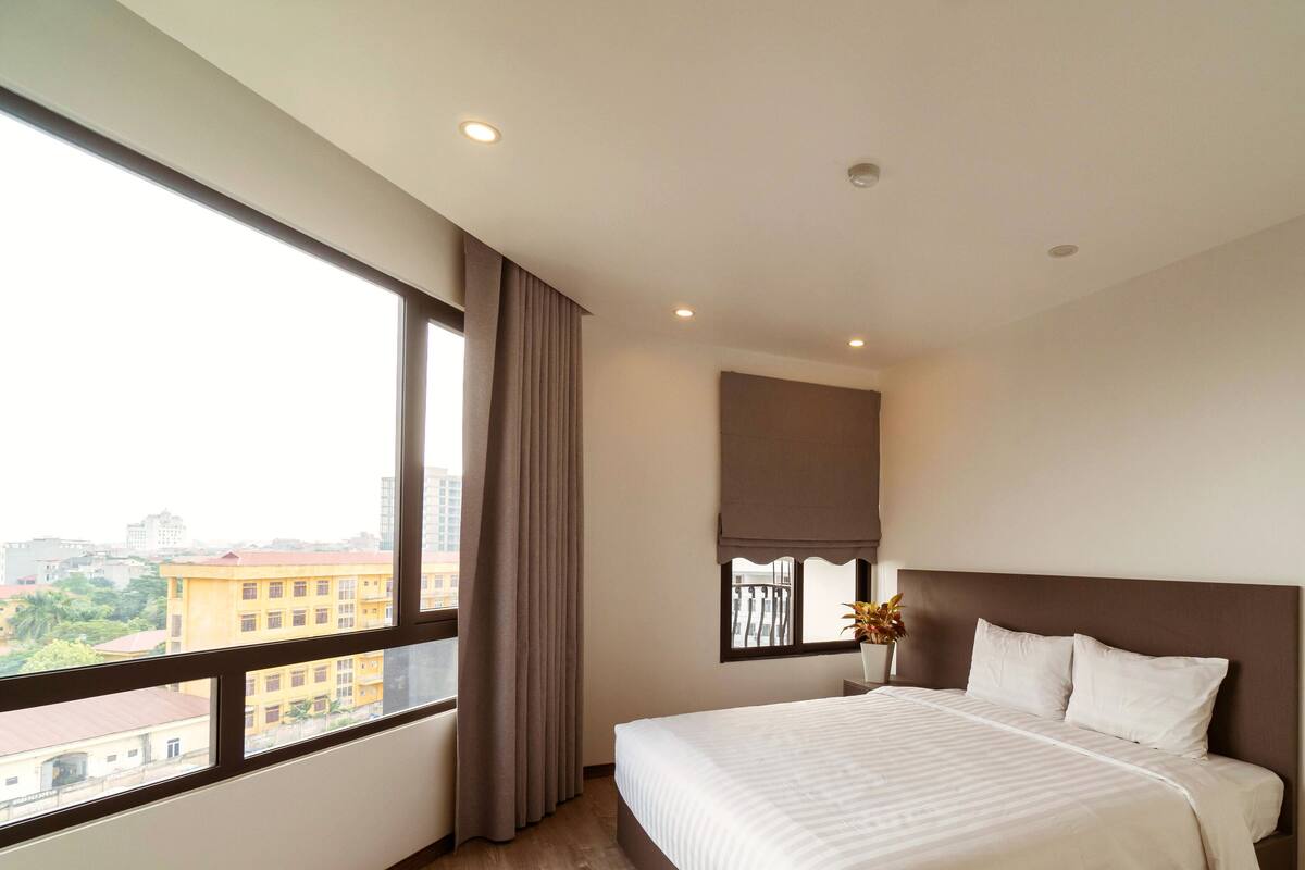 Une chambre d’hôtel avec une grande fenêtre, un lit avec du linge de lit blanc, une tête de lit marron et une plante en pot sur la table de chevet.