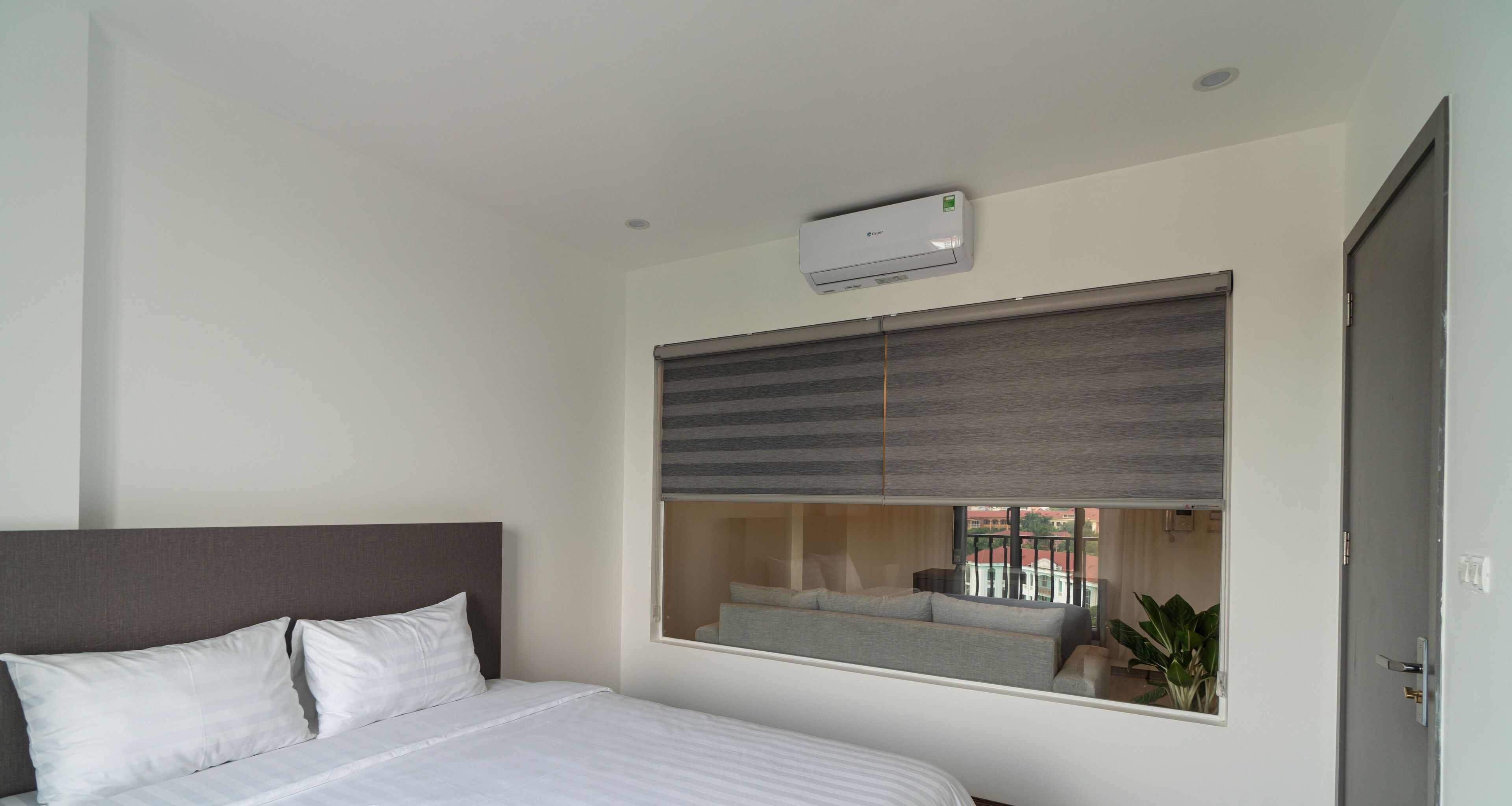 A modern hotel room with a large bed, a built-in air conditioning unit, a window with a view of the outside, and a door.