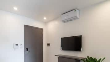 A modern, minimalist room with a flat-screen TV mounted on the wall, a built-in air conditioning unit, a wooden floor, a small table with two cups, a door, and a potted plant.