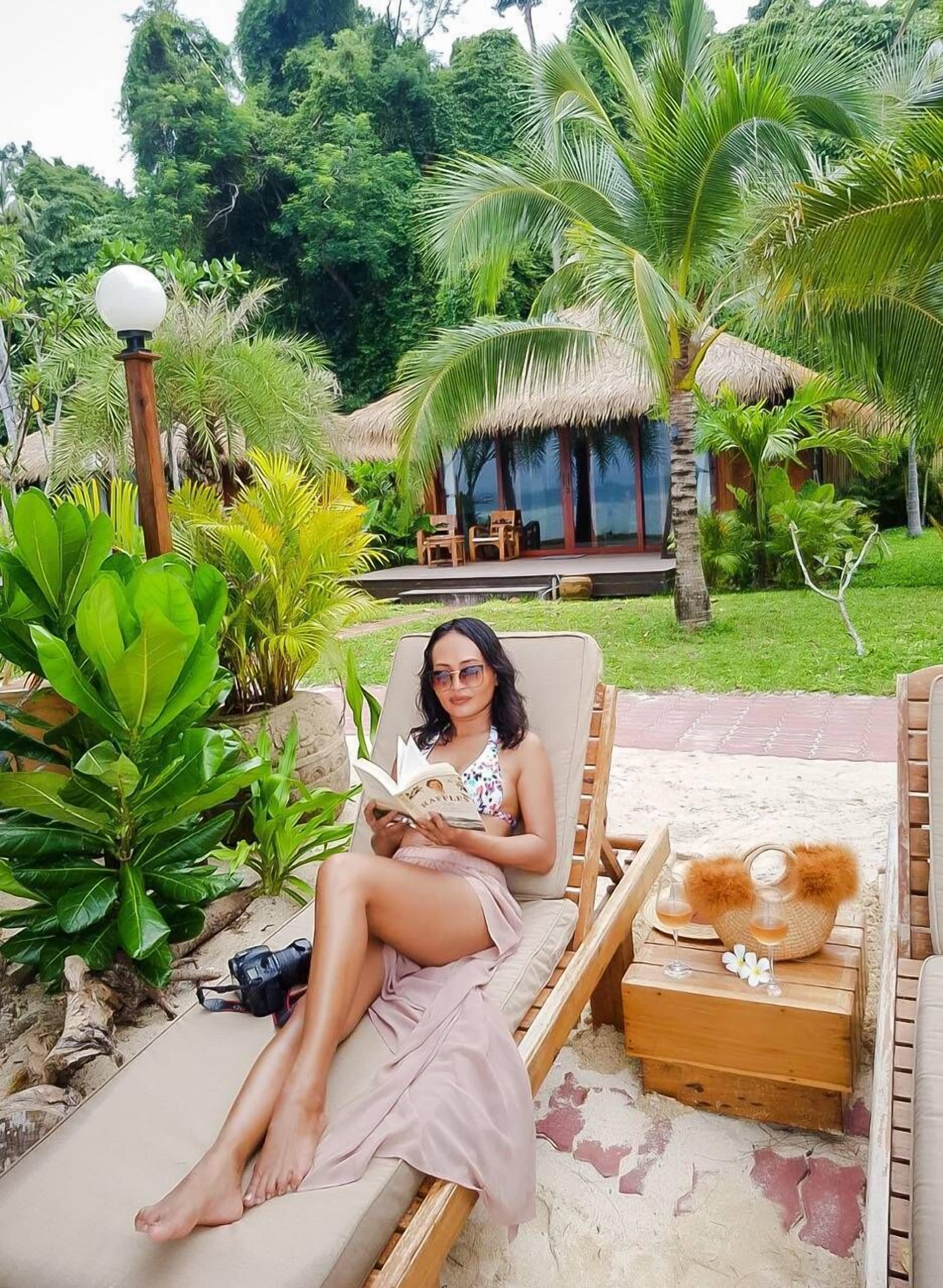 Une personne se détend sur une chaise longue dans un jardin tropical, avec une structure à toit de chaume en arrière-plan.