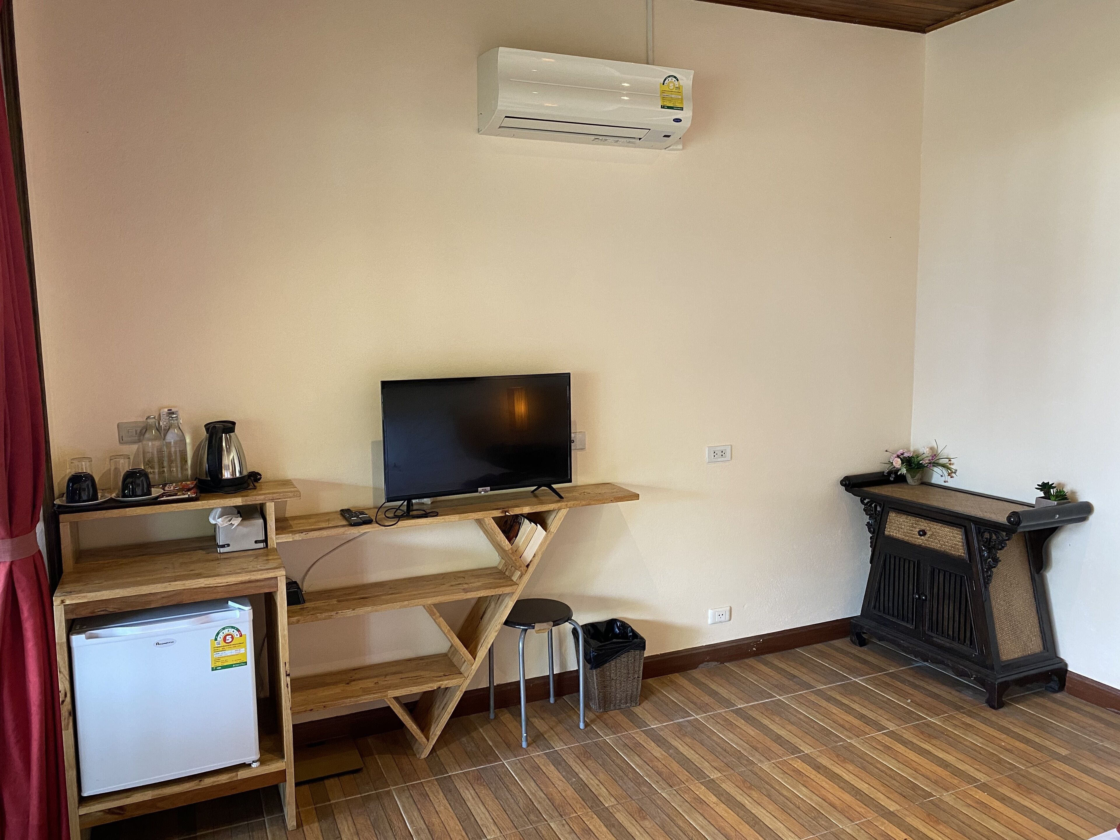 A compact living space with a television on a wooden stand, a small refrigerator, a trash bin, and a decorative console table.