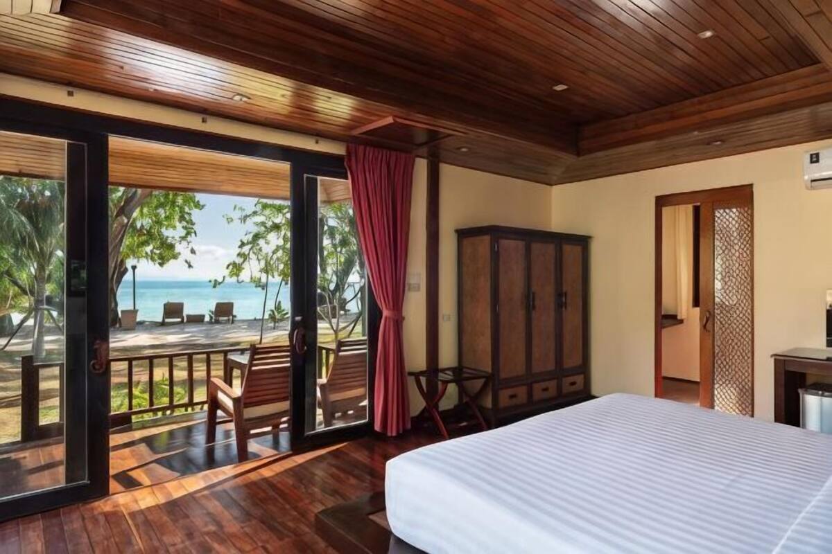 A bedroom with a large bed, wooden ceiling, a view of the beach, a wooden cabinet, and a door leading to another room.