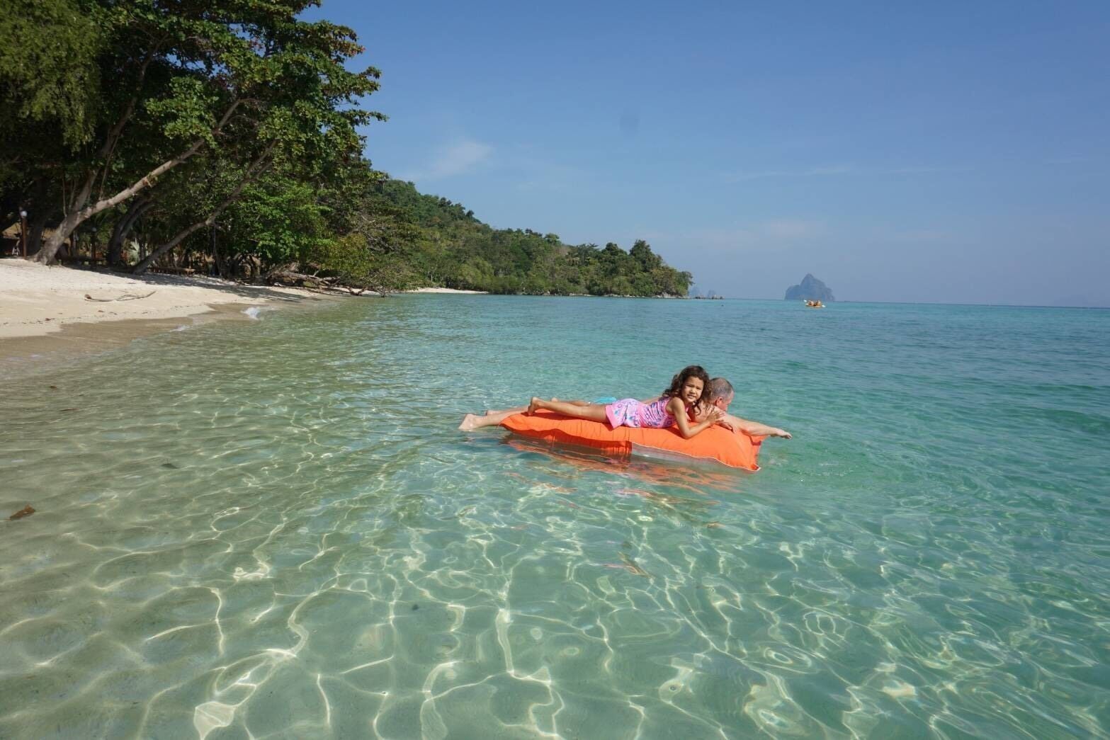A beach scene with clear water, a sandy shore, and a forested hill in the background.