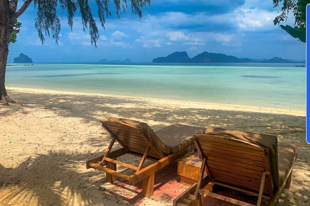 Une plage avec deux chaises longues en bois et une petite table en bois, offrant une vue sur une mer turquoise et limpide ainsi que sur des îles lointaines.