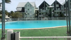 Uma piscina cercada por um muro branco, com edifícios ao redor que possuem varandas e telhado verde.