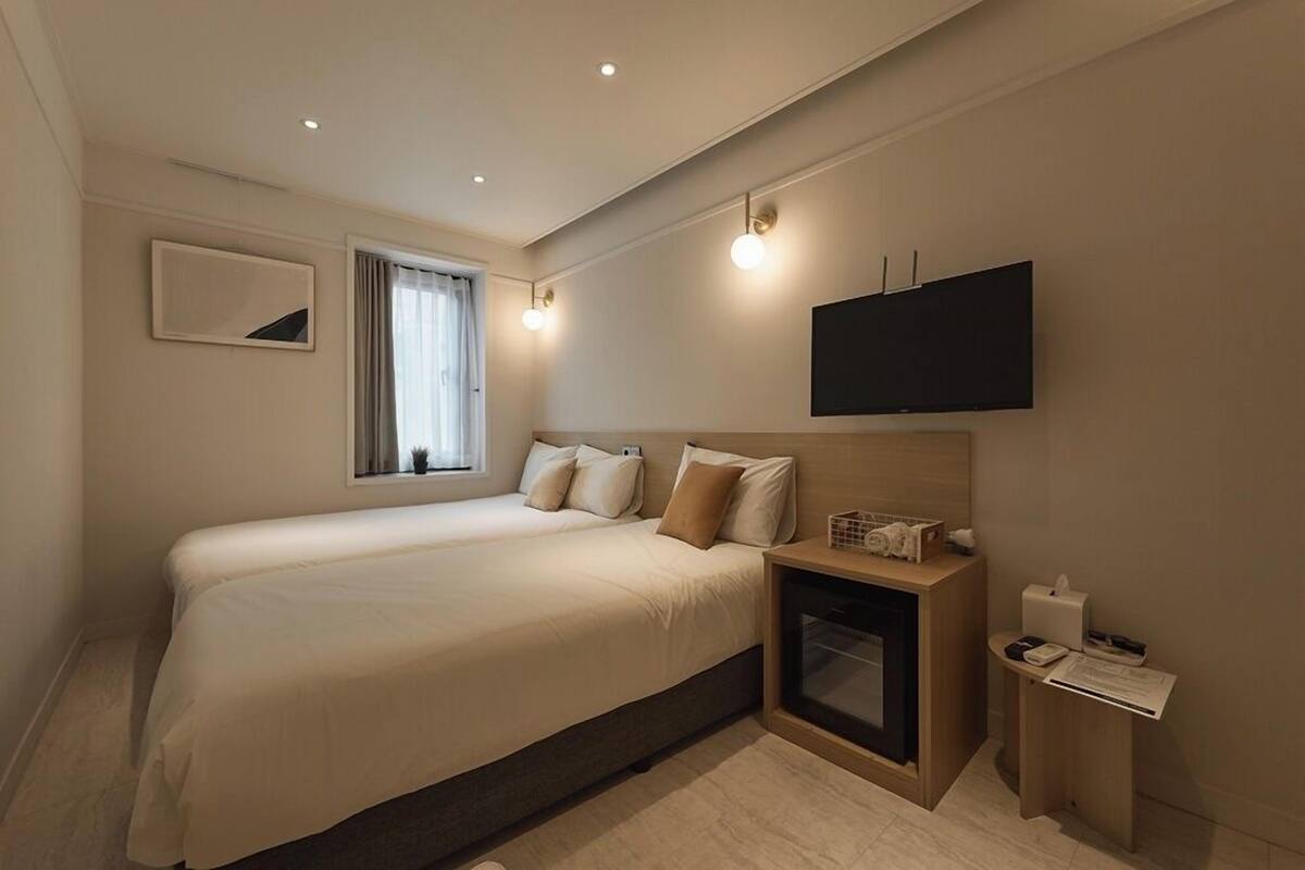 Habitación de hotel con una cama grande, una mesita de noche pequeña con lámpara, un televisor de pantalla plana en la pared y una ventana con cortinas.