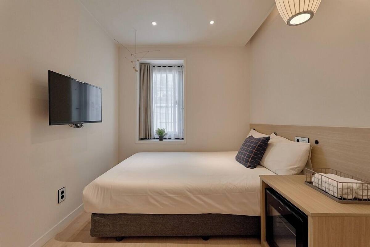 Un dormitorio moderno con cama, mesita de noche, televisor y ventana con cortinas.