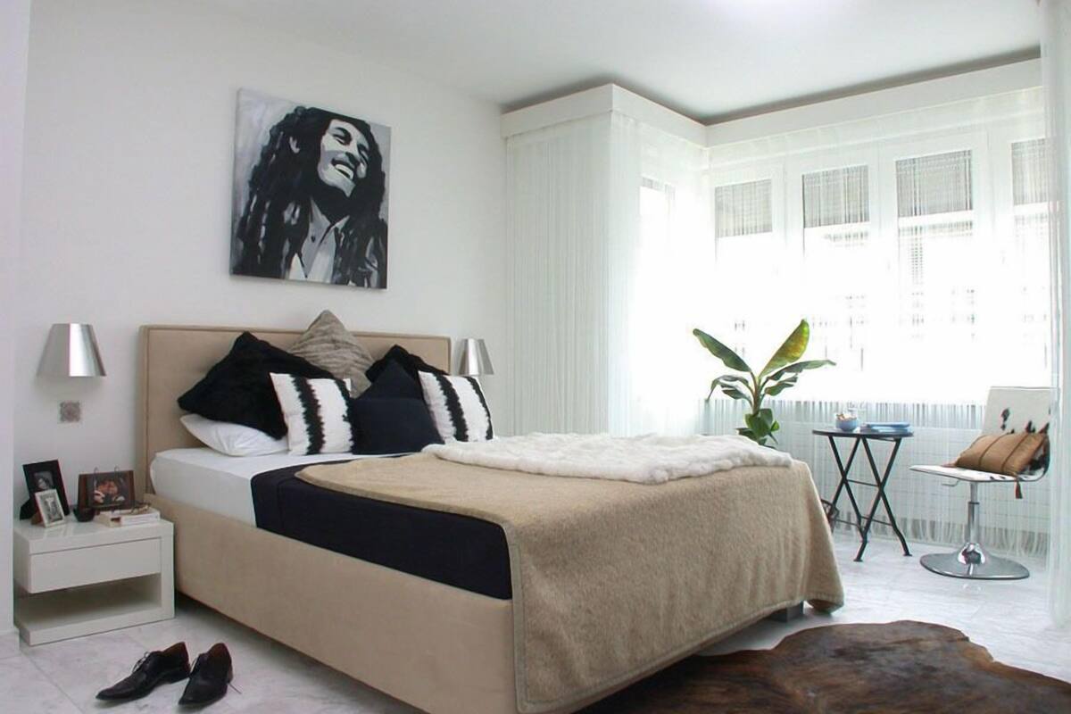A modern bedroom with a large bed, bedside tables, a small table with a plant, and a view of the city through large windows.