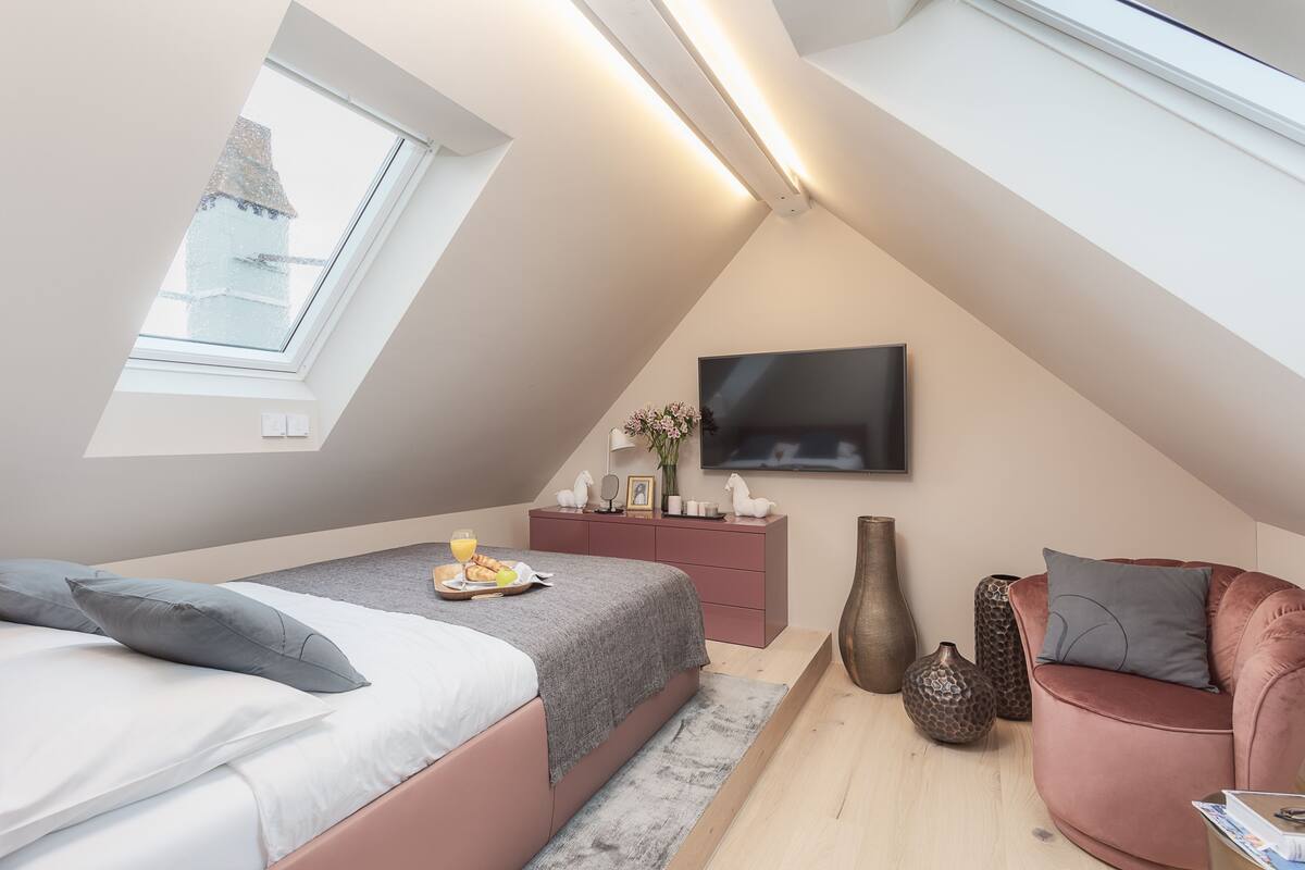 A bedroom with a sloped ceiling, a bed with pillows, a bedside table with a tray of food, a TV mounted on the wall, and a seating area with a chair and a sofa.