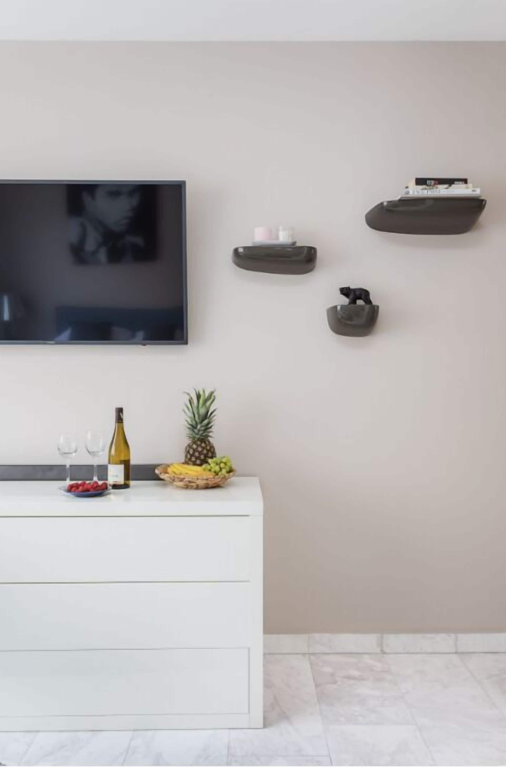 Ett modernt vardagsrum med en platt-TV som är monterad på väggen, ett vitt skåp med en vinflaska, glas och frukt, och två vägghyllor med dekorativa föremål.