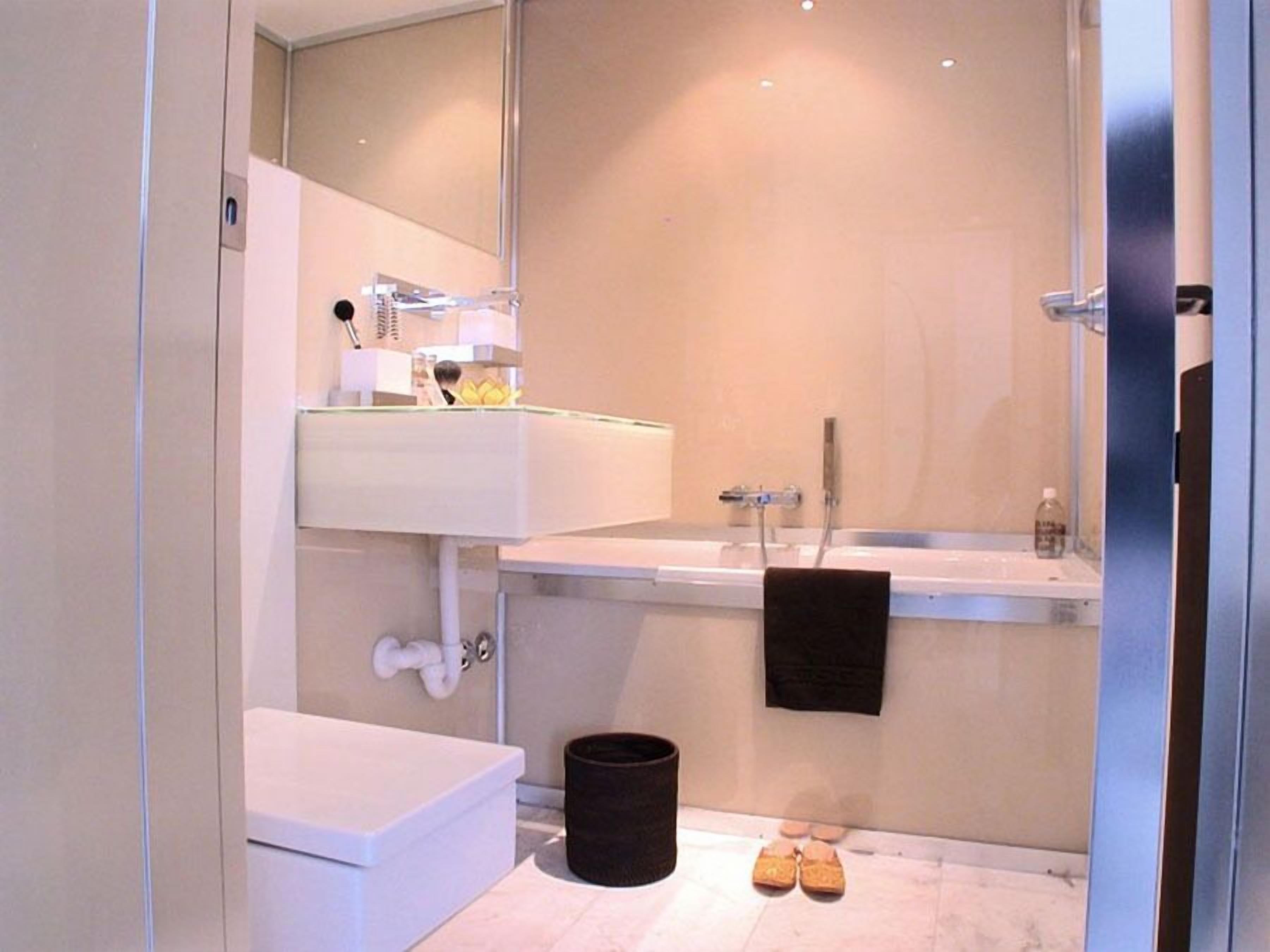 Une salle de bain moderne avec un lavabo blanc, un miroir et des toilettes. On y trouve une poubelle noire et une paire de chaussures sur le sol.