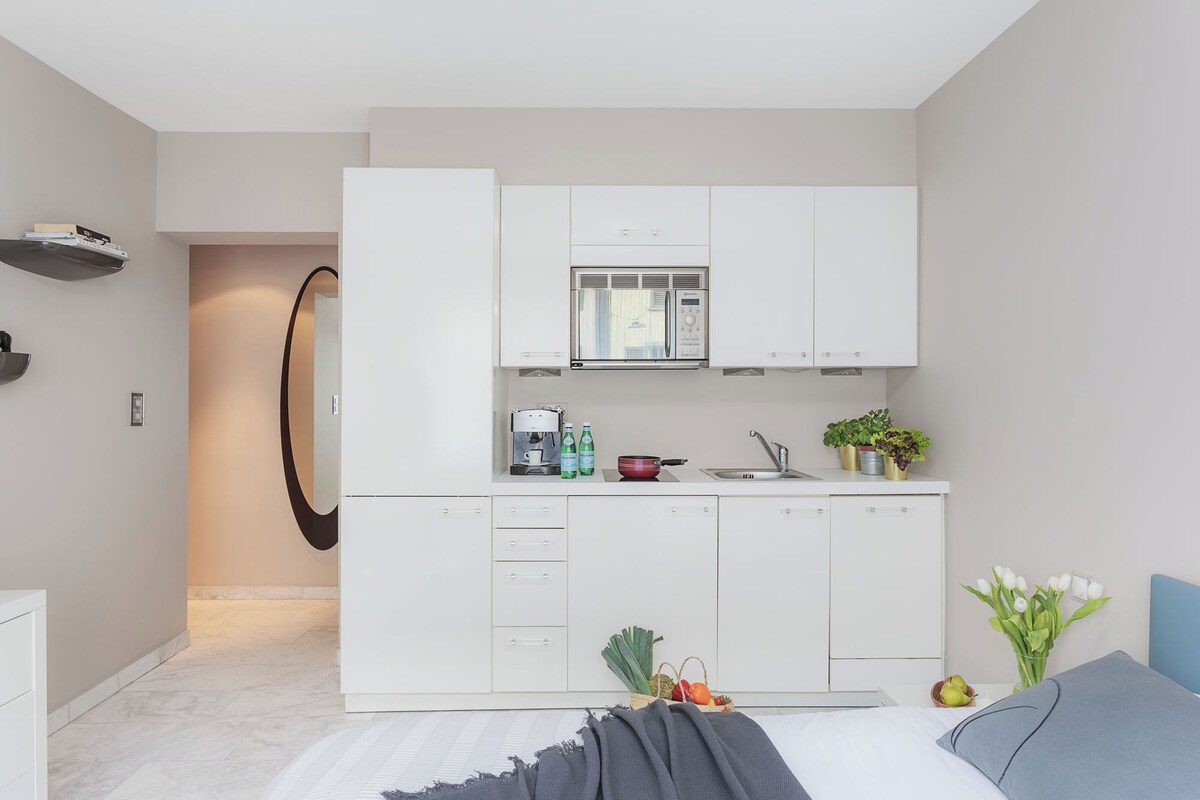 A modern kitchen with white cabinets, a microwave, and a sink. There is a bed with a blue pillow and a gray blanket.