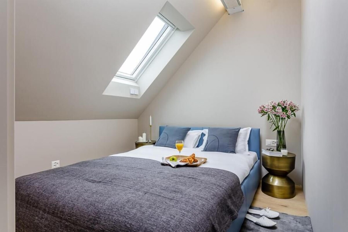 A bedroom with a skylight, a bed with a gray blanket, a bedside table with a vase of flowers, and a tray with breakfast.