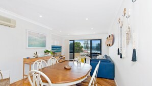 A modern dining area with a wooden table and white chairs, a blue sofa, and a large window offering a view of the outdoors.