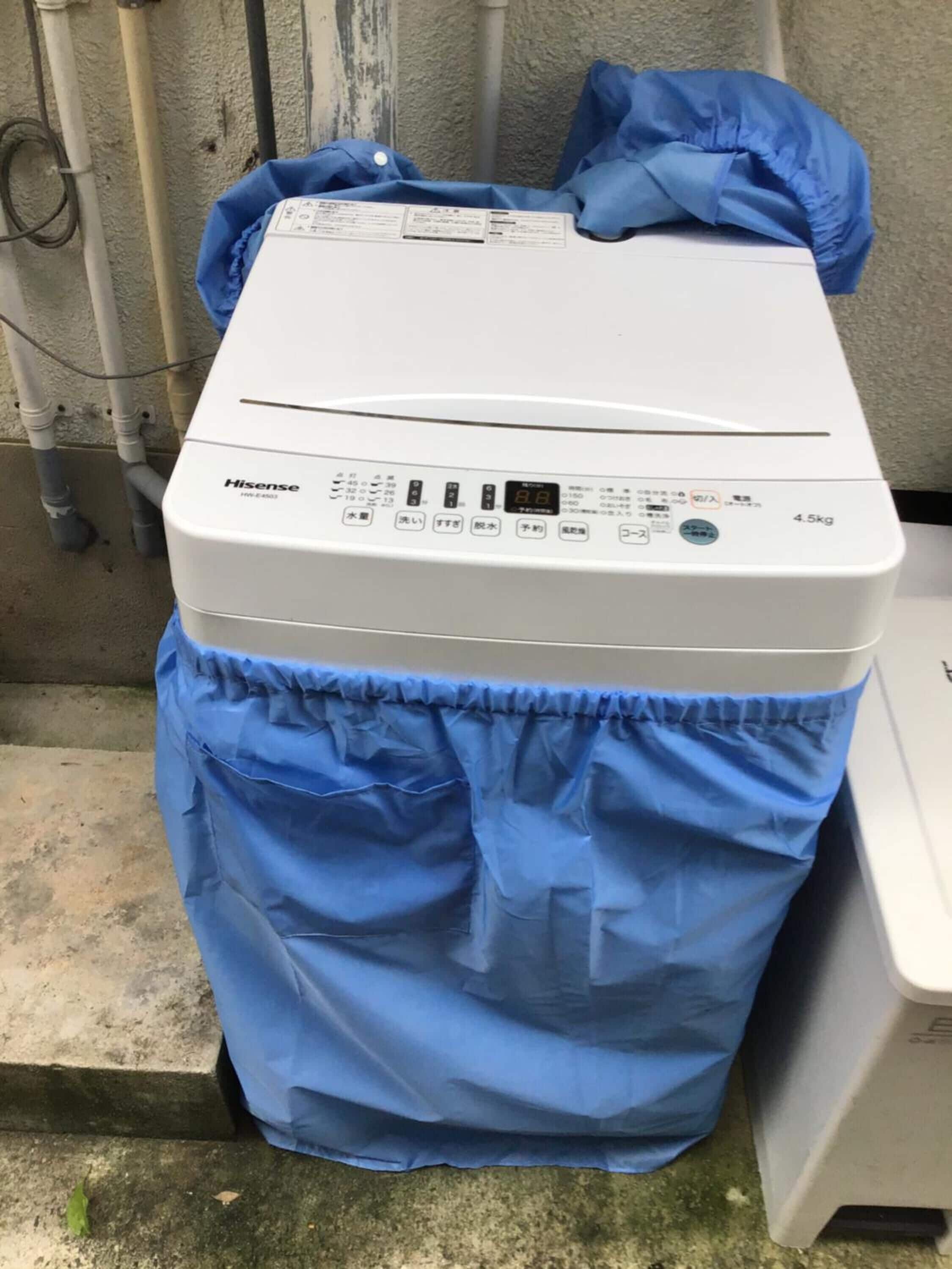 A white Hisense washing machine with a blue protective cover, placed outdoors near a wall with pipes.