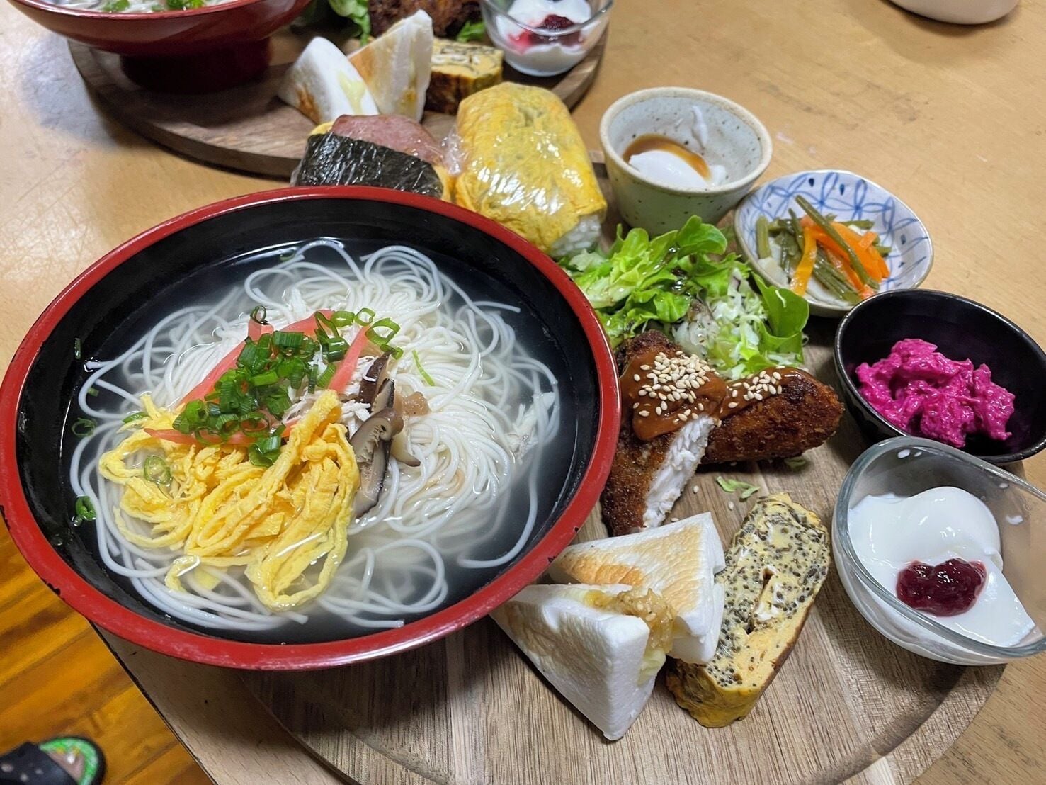 A variety of dishes including a bowl of noodles, sushi, and other side dishes on a wooden table.
