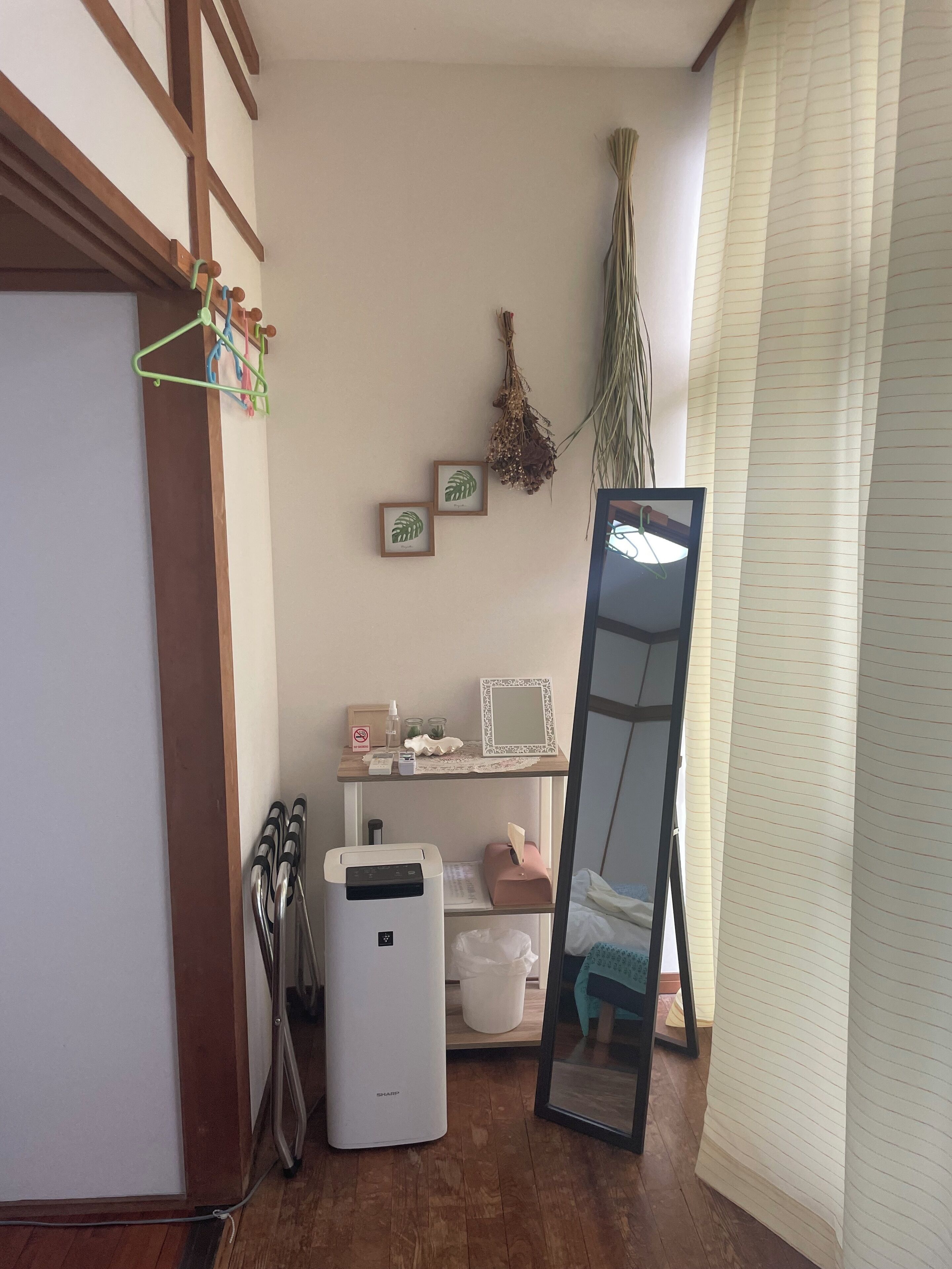 A room with a wooden floor, a white air purifier, a mirror, a shelf with toiletries, and a window with blinds.