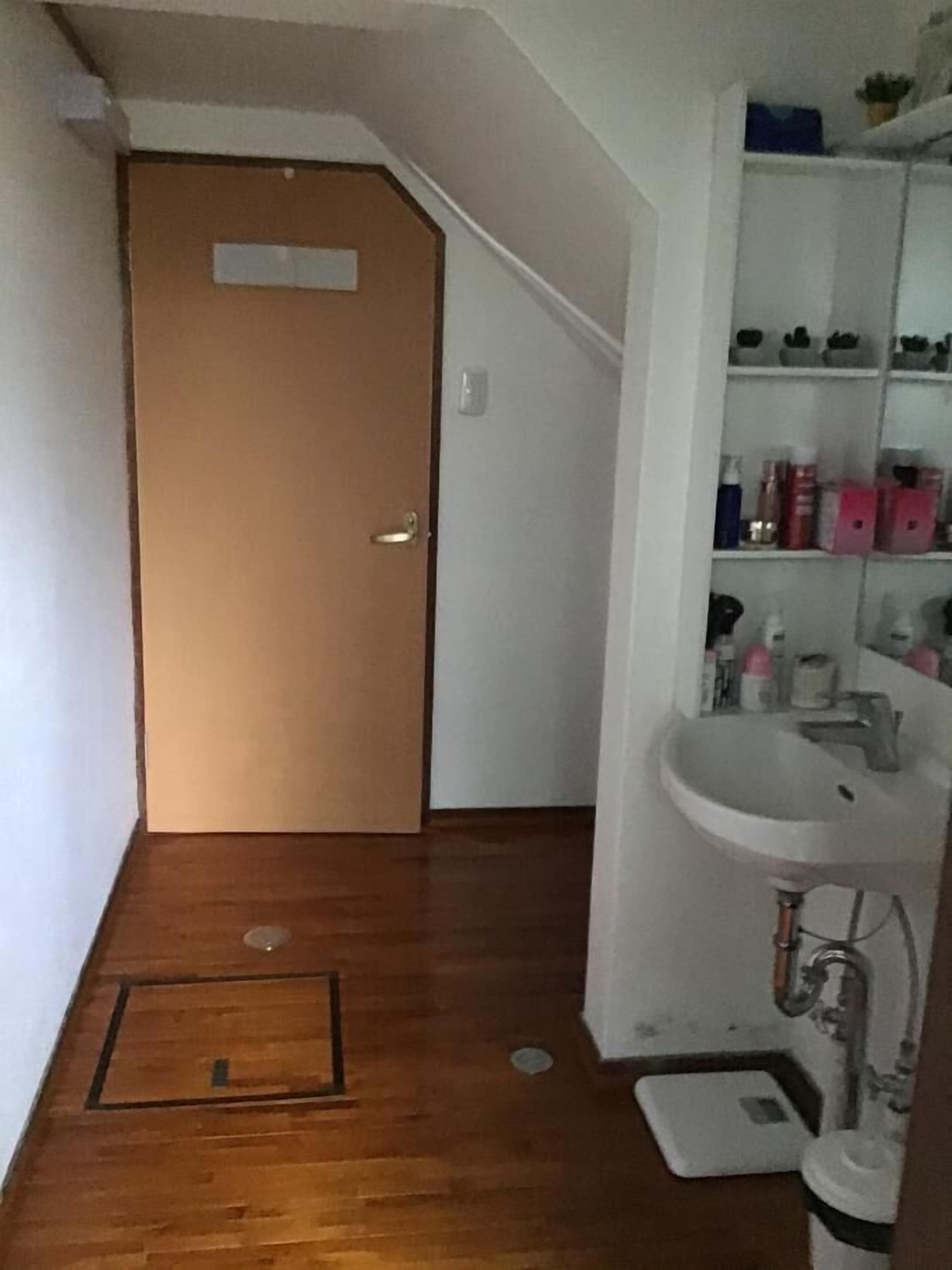A small bathroom with a wooden floor, a white sink, and a toilet. There is a door leading to another room.