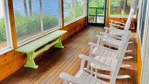 A wooden deck with white rocking chairs and a green bench overlooking a lake.