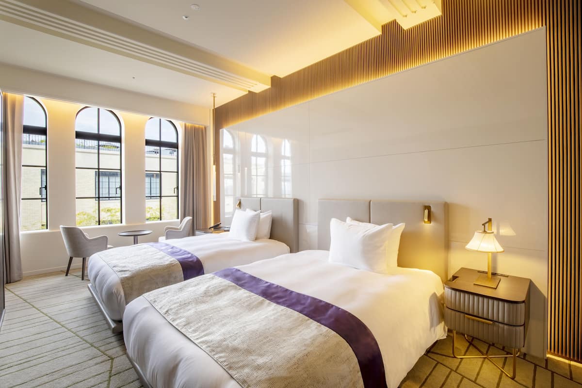 Une chambre d’hôtel avec deux lits, une petite table et une chaise. Elle est dotée de grandes fenêtres avec des rideaux, d’une table de chevet avec une lampe et d’un luminaire fixé au mur.
