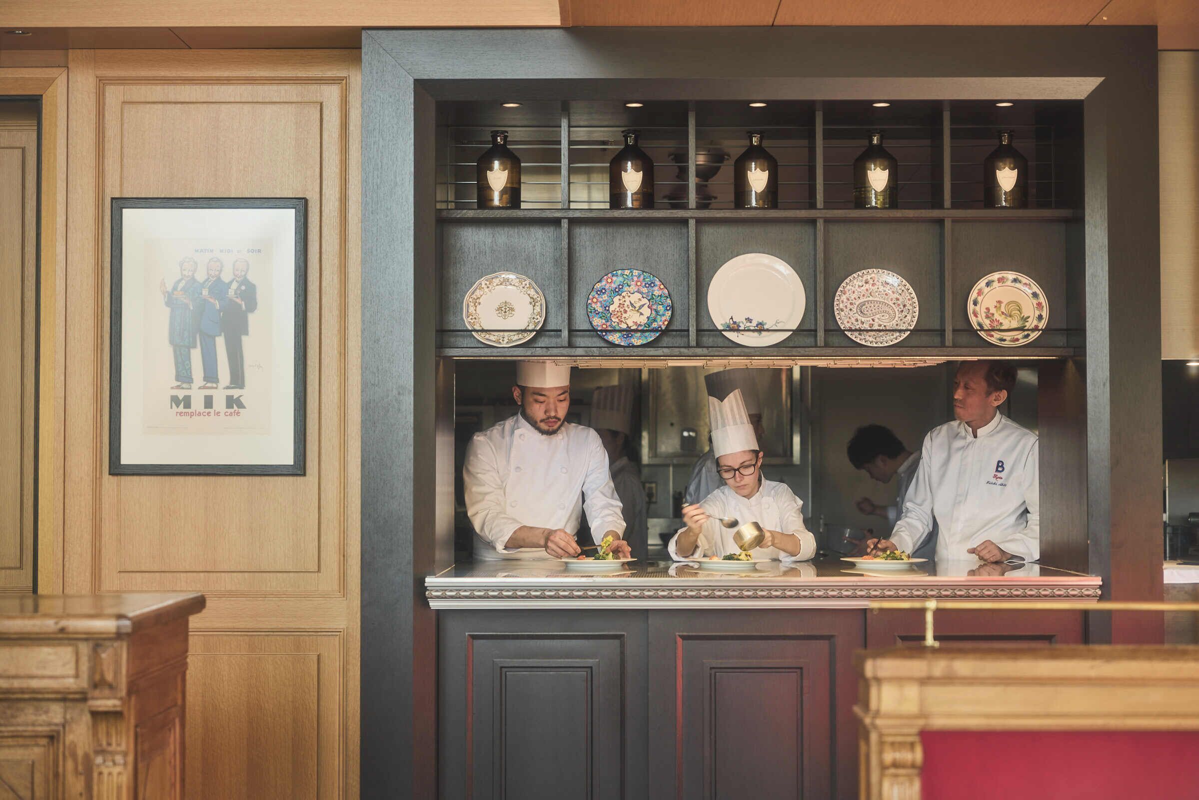 Une cuisine professionnelle où des chefs préparent des plats, une affiche encadrée accrochée au mur et des assiettes décoratives disposées sur des étagères.