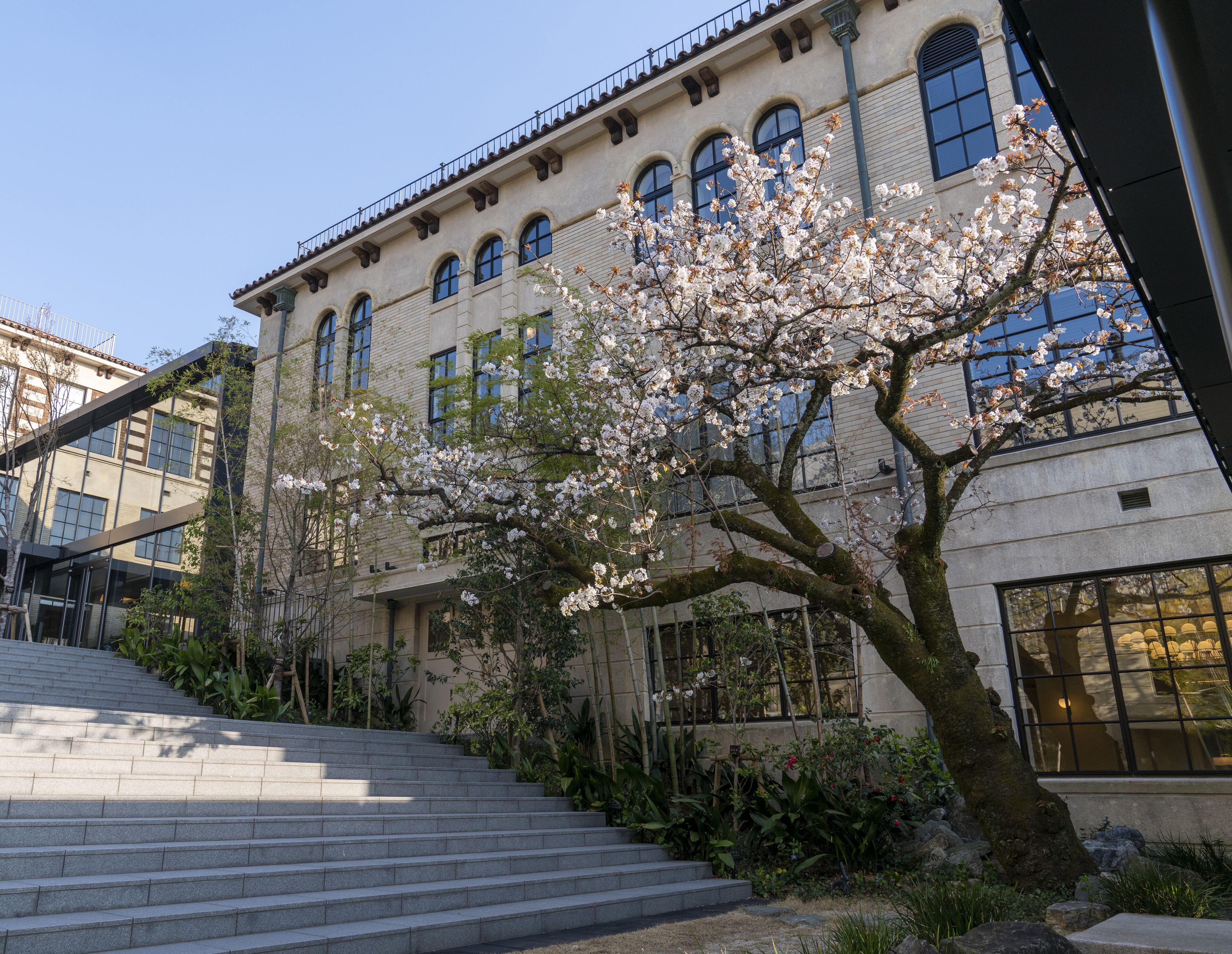A building with a staircase leading up to a balcony, surrounded by flowering trees and greenery.